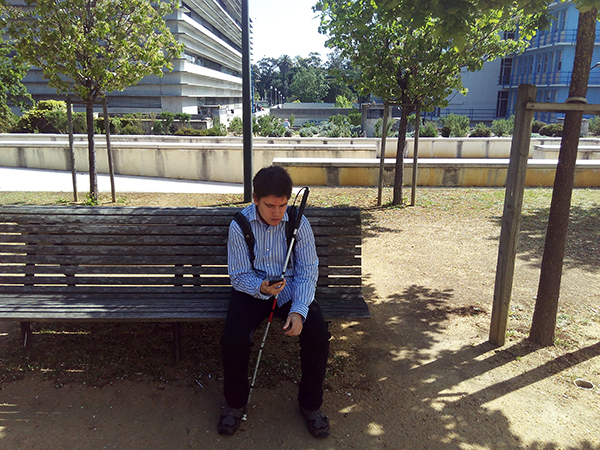 A blind person holding a white cane and seated on a park bench.