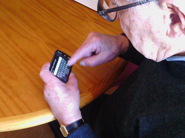 Older adult typing on a smartphone using a QWERTY keyboard. The person is using one hand to hold the phone and typing with a single index finger.
